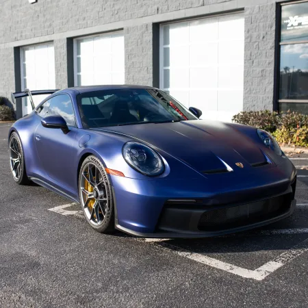 Matte blue Porsche sports car parked outside a grey building with white garage doors on a sunny day.