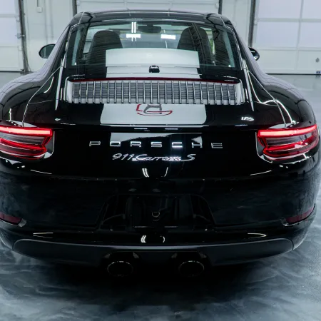 Rear view of a black Porsche 911 Carrera S in a spacious, polished indoor garage with bright lighting.