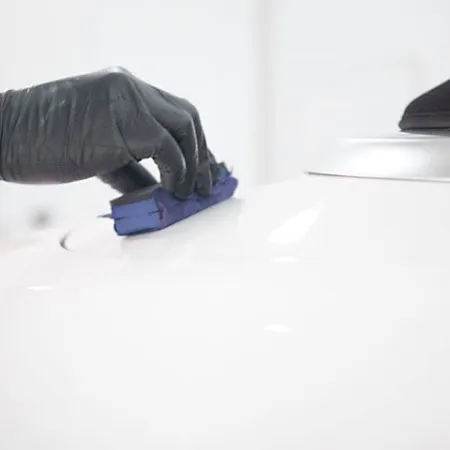 Person wearing black gloves polishing a white car surface with a blue sponge in a bright workshop.