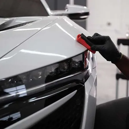 Person wearing black gloves detailing the front headlight and hood of a white sports car indoors.