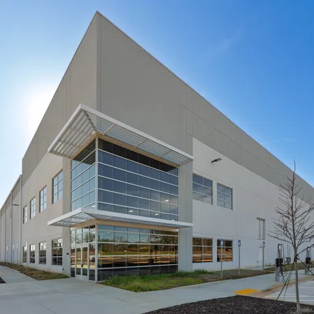 Large modern warehouse building with glass entrance under a clear blue sky and surrounding parking lot.