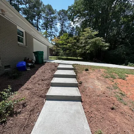Concrete steps leading up to a driveway beside a brick house with trees in the background under clear sky.