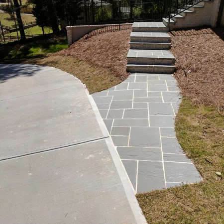 Stone pathway with stairs leading up to a house next to a concrete driveway in a landscaped yard.
