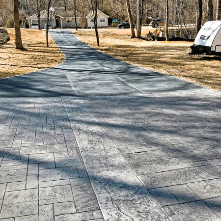 Long patterned concrete driveway leading to suburban home with RV and trees in a sunny yard.
