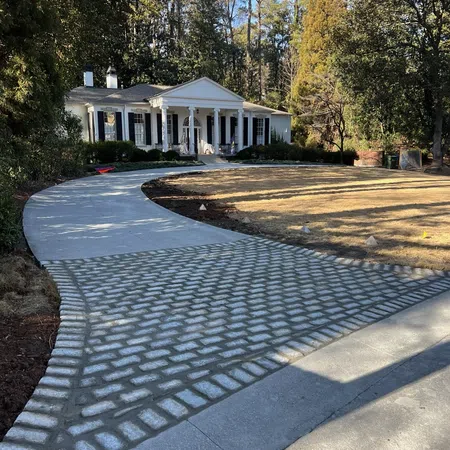 Curved concrete driveway with cobblestone section leading to white colonial house surrounded by trees in daylight