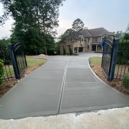 Newly poured concrete driveway leading to a large luxury house with iron gates and surrounding trees.