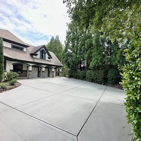 Spacious concrete driveway with basketball hoop beside a modern house surrounded by lush green trees and bushes.