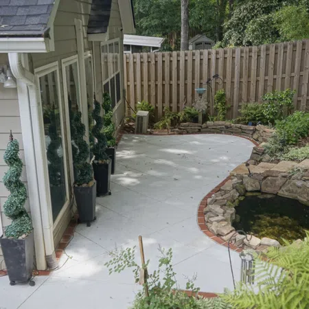 Residential backyard patio with shaped topiary, stone-bordered pond, and wooden privacy fence under trees.