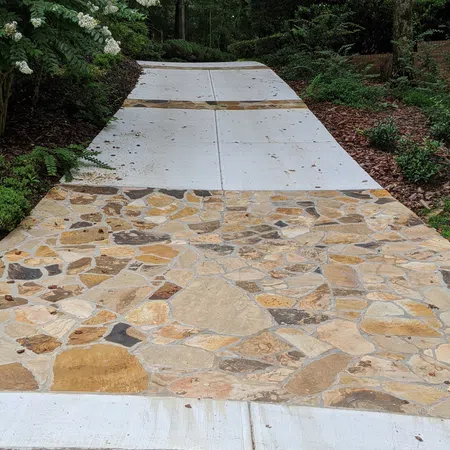 Driveway with mixed stone and concrete sections surrounded by green bushes and trees in a suburban setting.