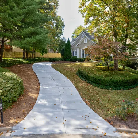 Curved concrete driveway leading to a charming house surrounded by trees and well-maintained landscaping.