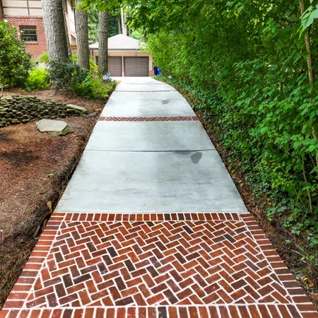 Concrete driveway with decorative brick inlays leading to a two-car garage surrounded by greenery and trees.