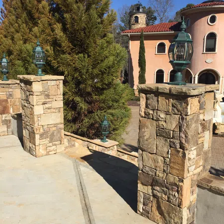 Stone pillars with vintage lanterns along a driveway leading to a Mediterranean-style house with trees under clear blue sky.