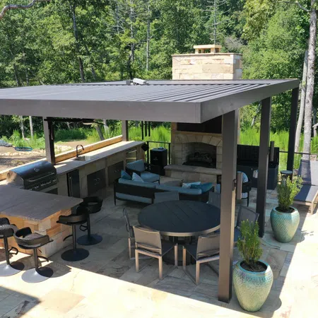 Modern outdoor patio with covered kitchen, bar stools, round dining table, fireplace, and lounge chairs surrounded by trees.
