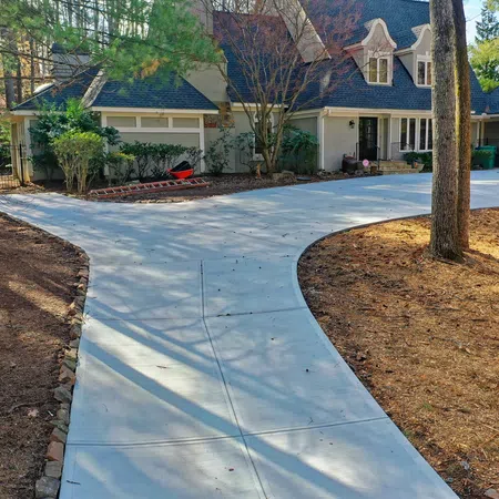 Curved concrete driveway leading to a large suburban house surrounded by trees and landscaped yard.