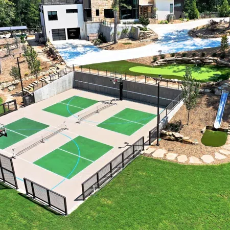 Outdoor multi-sport court with basketball hoops, pickleball nets, green turf, slides, and a modern house in the background