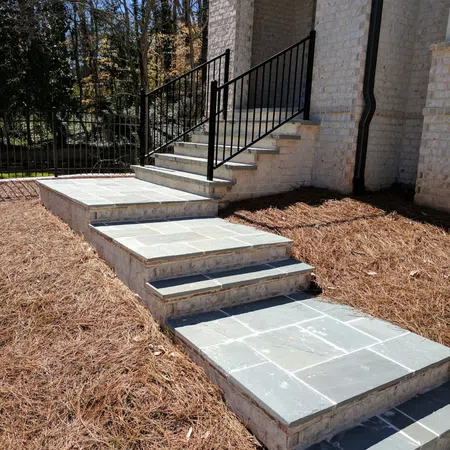 Stone steps with black metal railing leading up to white brick house entrance surrounded by pine straw mulch.