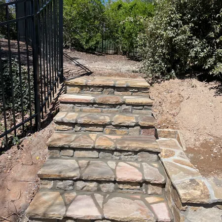Stone steps with irregular flagstone pattern leading uphill beside black metal fence and greenery under blue sky.