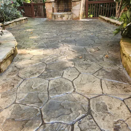 Outdoor stone patio with irregular patterned pavement and a stone fireplace surrounded by wood railing and greenery.