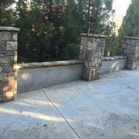 Stone pillars with low stone wall sections along a concrete driveway surrounded by greenery.