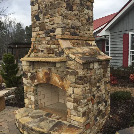 Outdoor stone fireplace with a curved hearth and brick interior on a patio with surrounding landscaping.