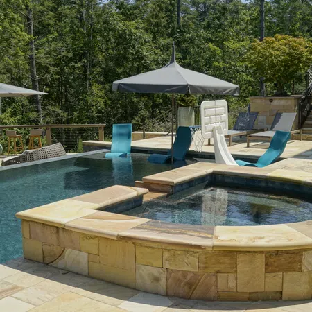 Luxury outdoor pool area with built-in hot tub, lounge chairs, umbrellas, and surrounding forest scenery.