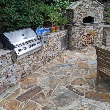 Outdoor stone kitchen with stainless steel grill, stone fireplace, potted plants, and natural stone patio flooring.