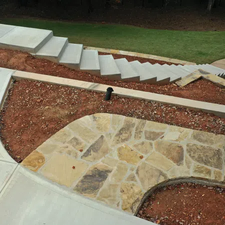 Outdoor stone patio with surrounding bare soil, concrete walkway, and stairway in a garden setting.