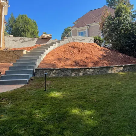 Concrete stairs lead up a landscaped hill with red soil, retaining wall, adjacent house, and green grass under blue sky.