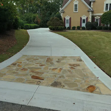 Curved driveway with a stone inlay leading to a beige house surrounded by green lawn and trees.