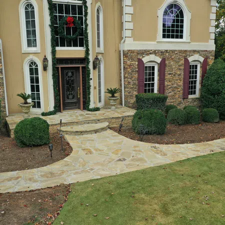 Stone pathway leads to elegant house entrance with wreath, stone walls, arched windows, and manicured shrubs.