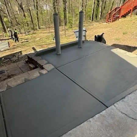Freshly poured concrete slab with drainage pipes, surrounded by wooded area and a wooden deck in the background.