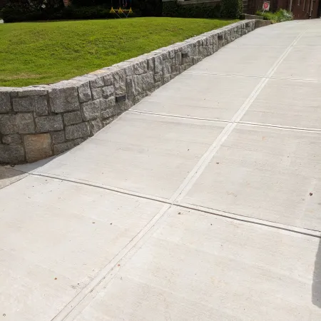 Smooth concrete driveway with stone retaining wall and green lawn beside a brick house with flower pots.