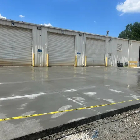 Newly poured concrete slab outside warehouse with three closed loading dock doors and caution tape.