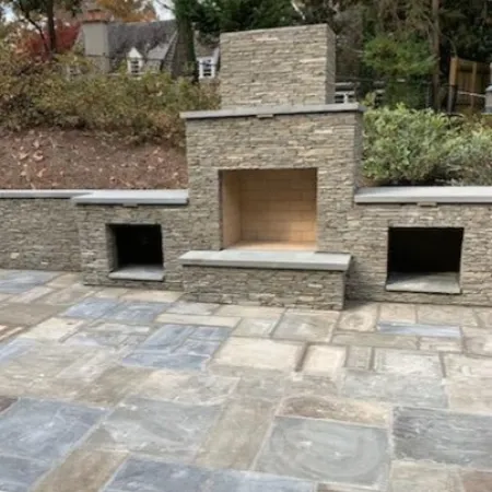 Outdoor stone fireplace setup surrounded by landscaped patio and greenery.