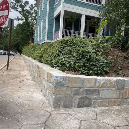 Stone retaining wall with green ivy plants beside blue house with porch and sidewalk on street corner.
