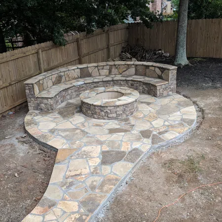 Stone patio with curved stone bench and round fire pit in a backyard surrounded by wooden fence.
