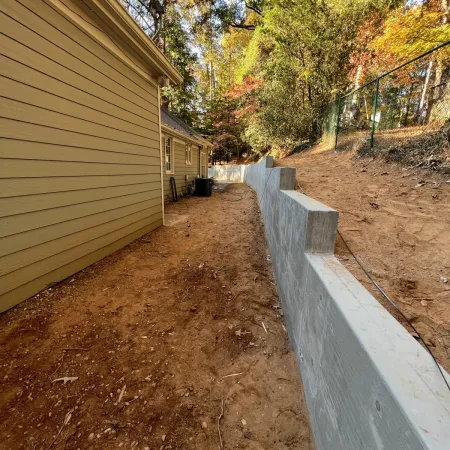 New concrete retaining wall runs alongside yellow house with bare dirt and autumn trees in background.