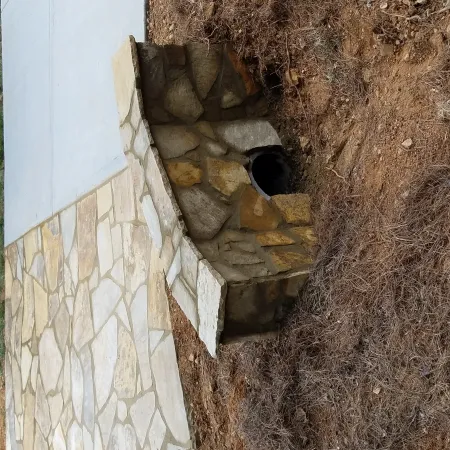 Stone culvert with pipe under concrete sidewalk surrounded by dry pine needles and soil.