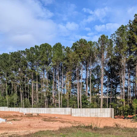 Concrete retaining wall under construction on dirt land with tall pine trees and blue sky in the background