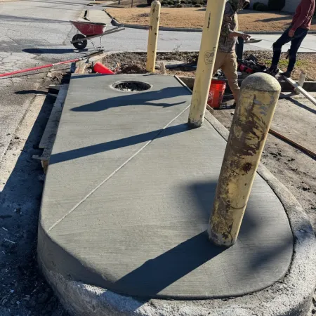 Newly poured concrete island with yellow bollards and workers preparing the area on a sunny day.