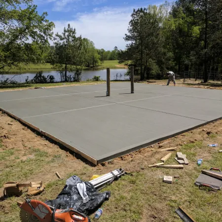 Freshly poured concrete volleyball court with posts near a lake and surrounded by trees on a sunny day.