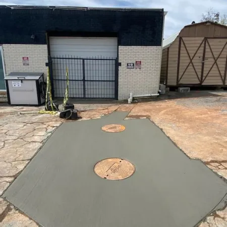 Freshly poured concrete with two circular manhole covers in a parking lot setting, surrounded by storage buildings.