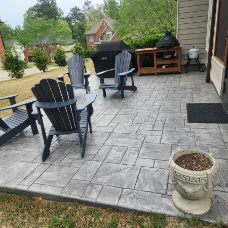Stylish outdoor patio featuring gray stamped concrete, Adirondack chairs, and a BBQ grill, surrounded by greenery.