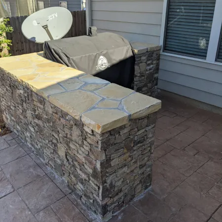 Outdoor stone countertop and grill area with stamped concrete patio and beige house siding in daylight.