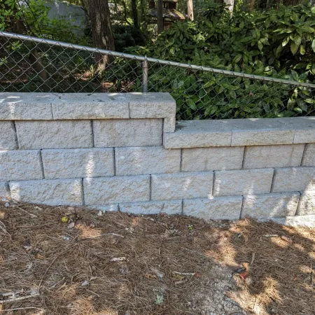 Gray concrete block retaining wall with stepped design beside a chain link fence and pine needle ground cover