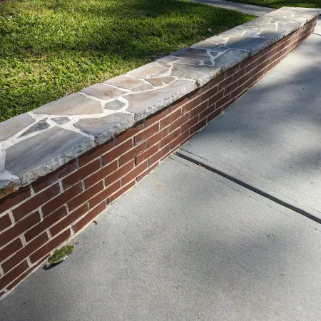 Brick planter wall topped with irregular stone slabs beside green grass and concrete sidewalk on sunny day.