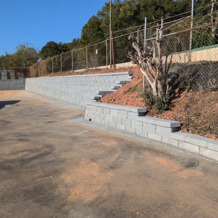 Concrete retaining walls with steps lining a dirt slope beside an asphalt area under clear blue sky.