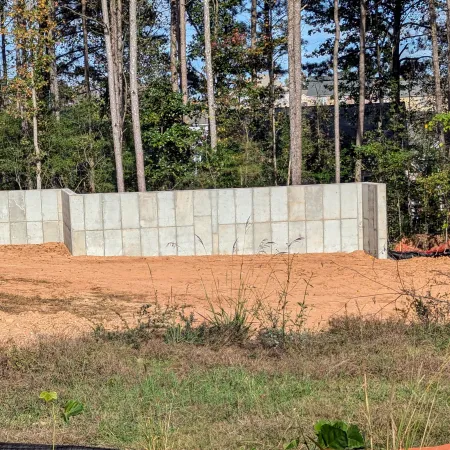 Concrete retaining wall with bare earth and grassy area in front, surrounded by tall trees under blue sky.