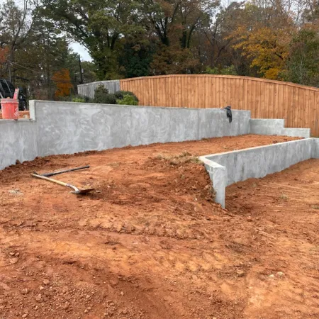 Concrete retaining walls surround freshly excavated red soil with tools nearby in a residential backyard.