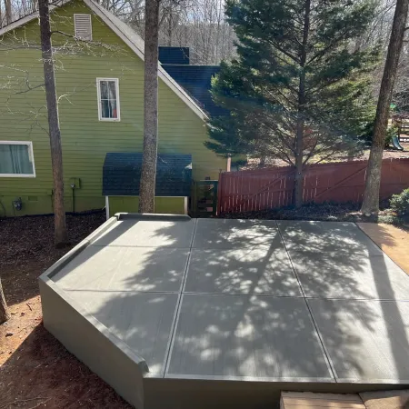 Newly poured concrete slab patio in backyard with trees, green house, and red wooden fence in background.